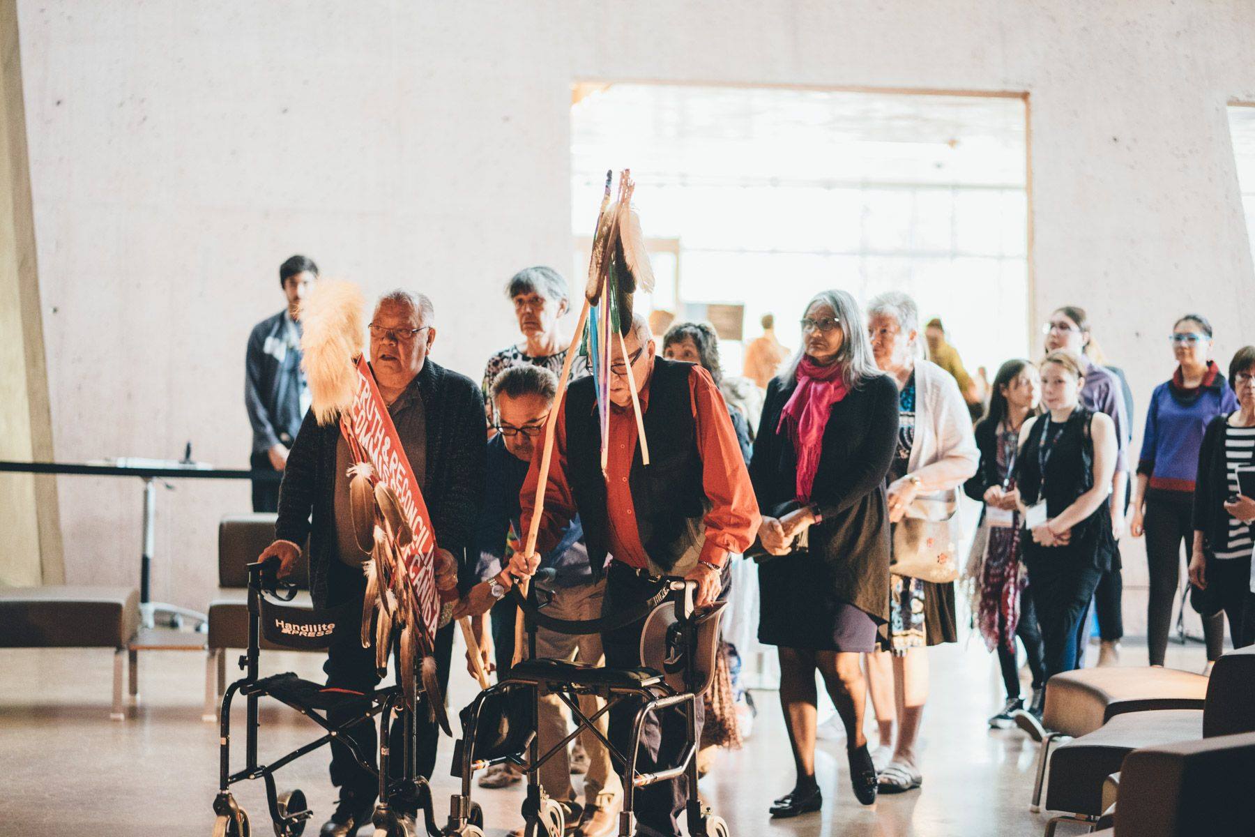 A procession of Survivors and people at an Imagine a Canada event, some with walkers. One man is holding the Eagle Staff.