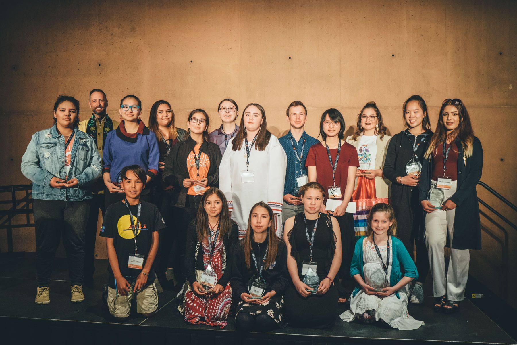 A group of 17 students and teachers pose in front of a light brown backdrop at an Imagine a Canada event.