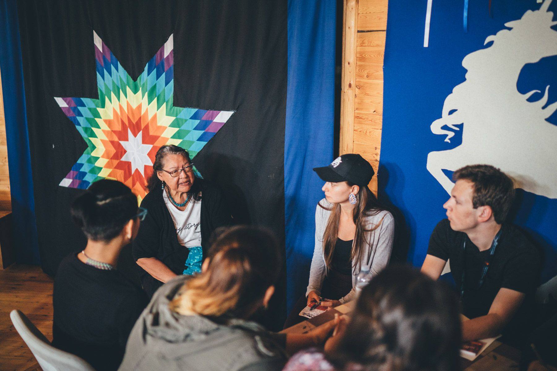 A small circle of people listen to Elder Paynter in front of a black quilt with a bright Indigenous multi-coloured star.