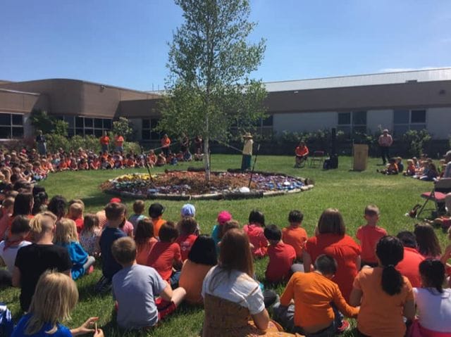 A large group of youths sitting in a circle around a tree. Many are wearing orange shirts, and one person stands by the tree.