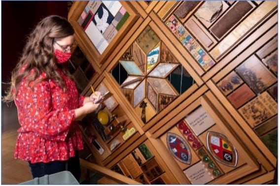 Woman writing on small pad standing in front of art installation made with wood and mixed materials