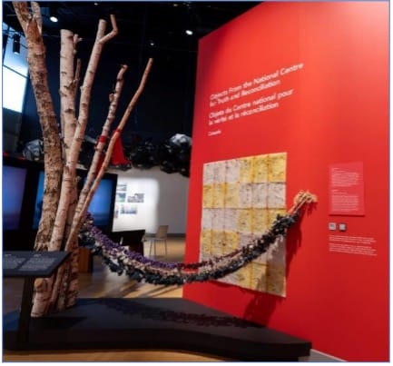 Image of art installation by Cree artist Linda Young of Saskatoon representing an empty baby swing (wêwêpison), strung from a birch tree to the wall of the installation. The swing is affixed with dozens of tobacco pouches