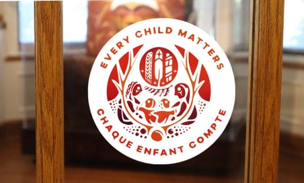 Product image of window decal with Survivors' Flag and text "every child matters" and "chaque enfant compte" on a glass pane of a wooden framed door with the bentwood box in the background.