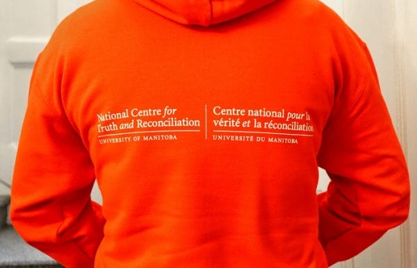 Product image of an orange hooded sweatshirt with white text "National Centre for Truth and Reconciliation" over "University of Manitoba" and "Centre national pour la vérité et la réconciliation" over "université du manitoba".