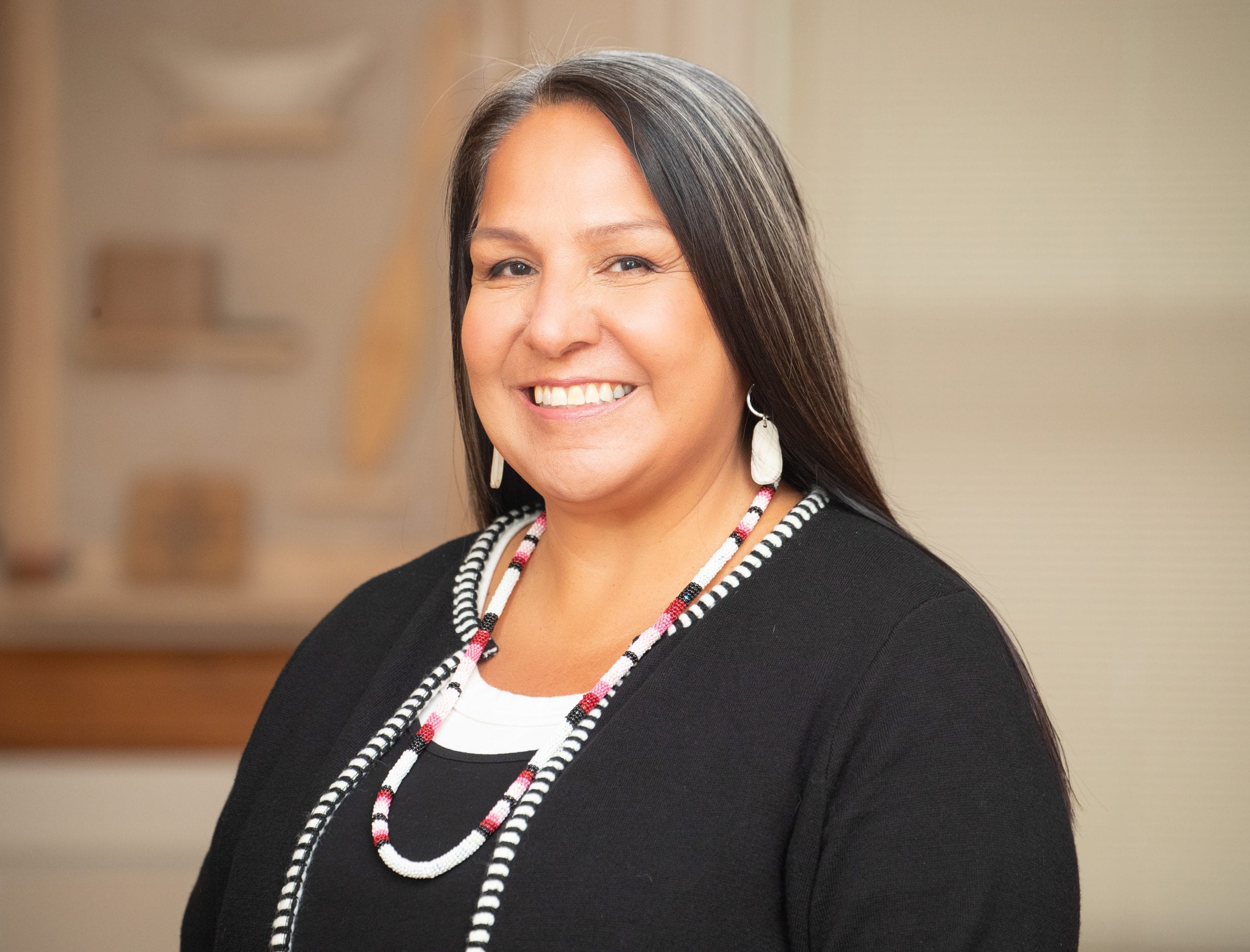 Stephanie Scott has long, straight salt and pepper hair and is wearing earrings, a beaded necklace and a black cardigan over a black and white shirt.