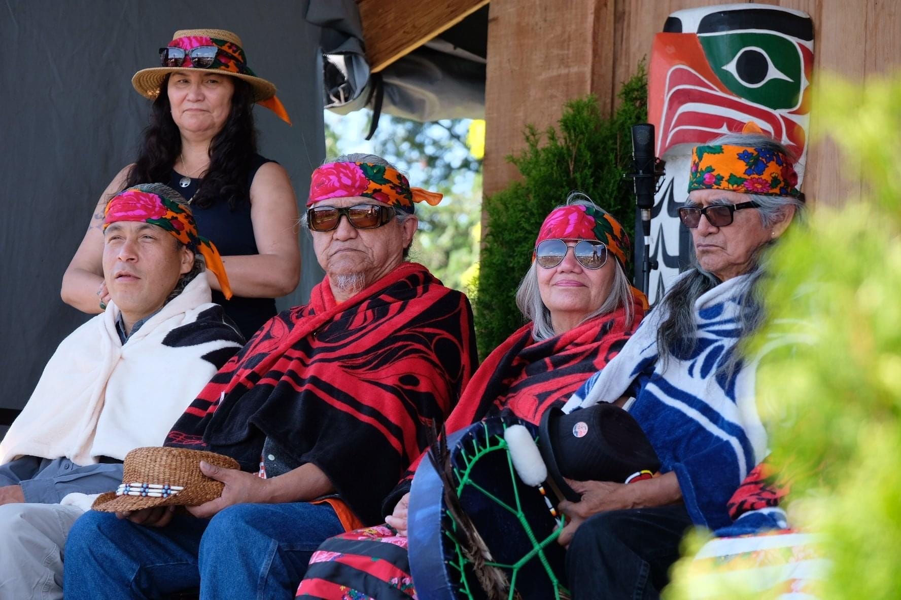 A group of Indigenous individuals are seated outdoors, wearing patterned blankets and floral headbands.