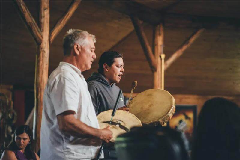 Two men play frame drums in a lodge.